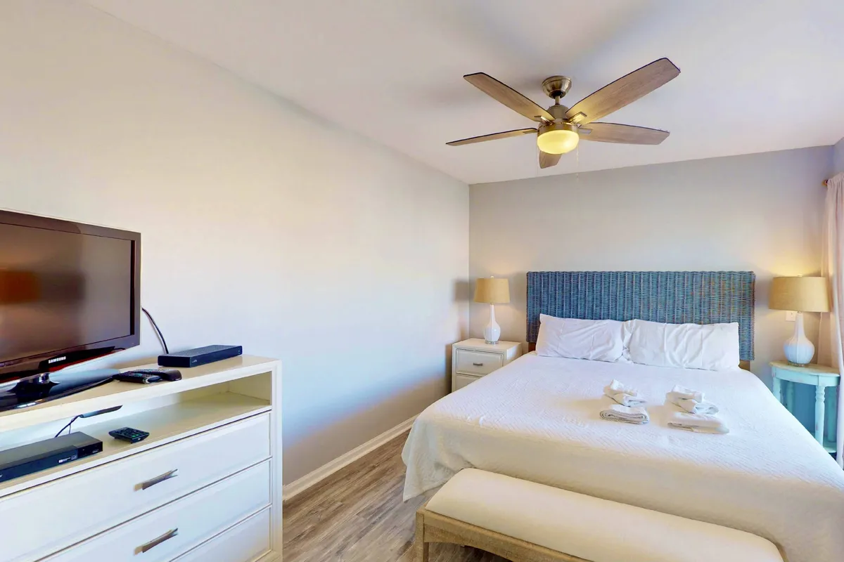 A calm and inviting bedroom with a bed adorned with decorative pillows, a ceiling fan above, a television on a dresser, and side tables with lamps. Natural light streams in through a window, contributing to a relaxing, restful mood.
