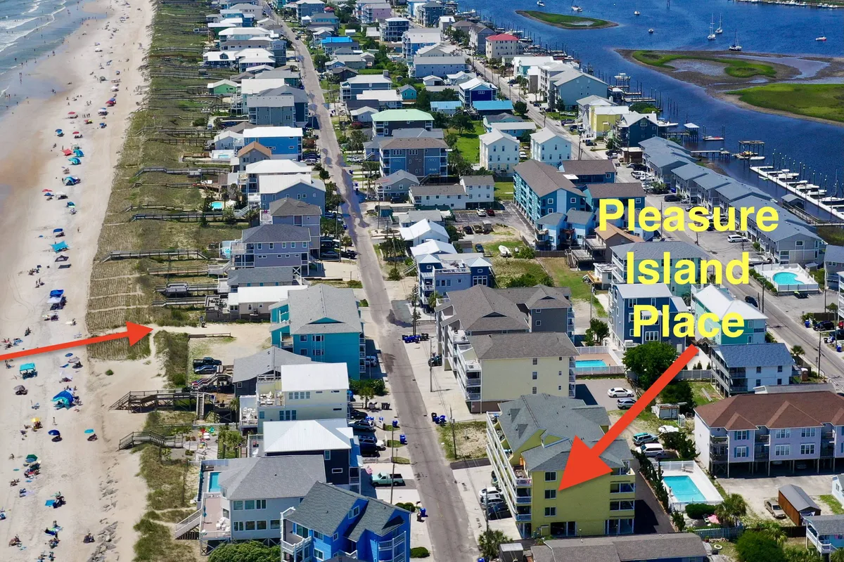 The image features the Pleasure Island Place building with a clear view of the beach access path, surrounding houses, and the ocean in the background. The coastline is visible, hinting at the proximity to both the sand and local attractions.