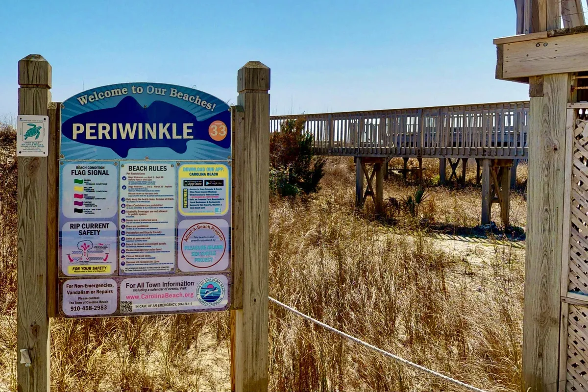 A welcoming scene with a 'Welcome' sign, a wooden boardwalk leading toward the beach, and beach grass swaying in the breeze. The beach and ocean are visible, inviting exploration and adventure.