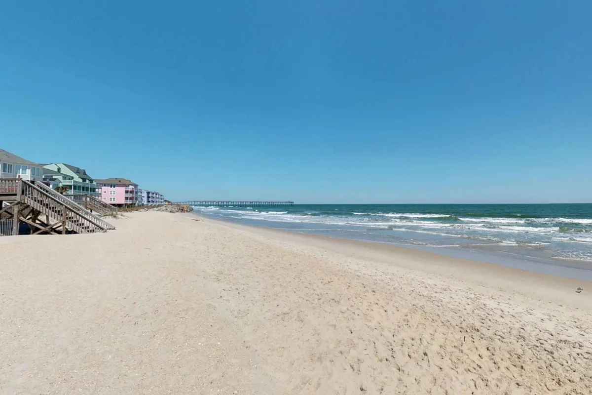 A wide view of the beach and ocean, with a pier stretching into the water and colorful houses lining the shoreline. The area is lively but not overcrowded, suggesting opportunities for beach walks, sunbathing, and exploring vibrant local life.