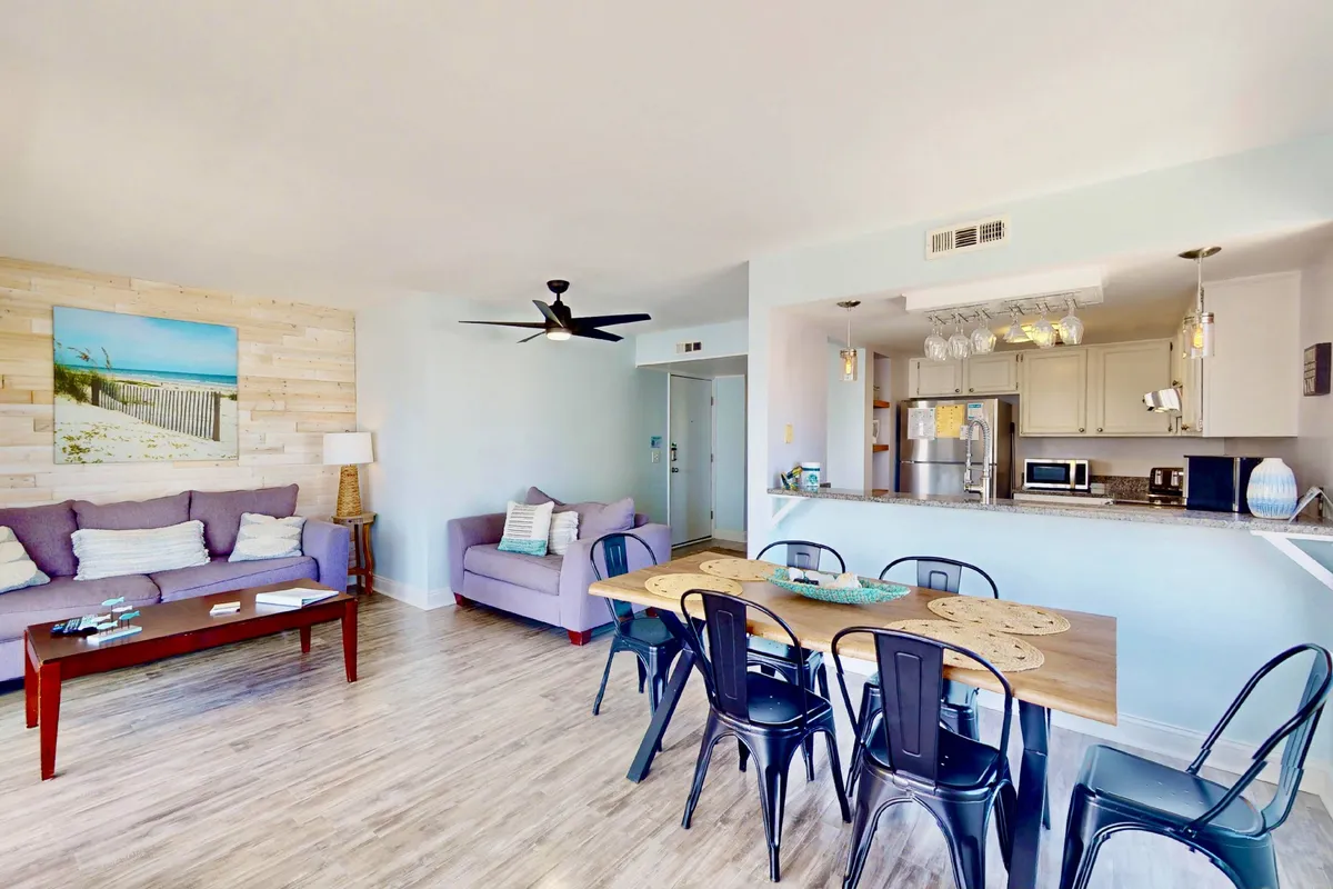 This image captures a spacious, open-plan interior with a modern kitchen, dining table, and a living area featuring a cozy purple couch. The kitchen area is equipped with modern appliances and is visually connected to the dining and living spaces, emphasizing togetherness and ease of entertaining. Beach-themed artwork adds a coastal touch, reinforcing the vacation vibe.