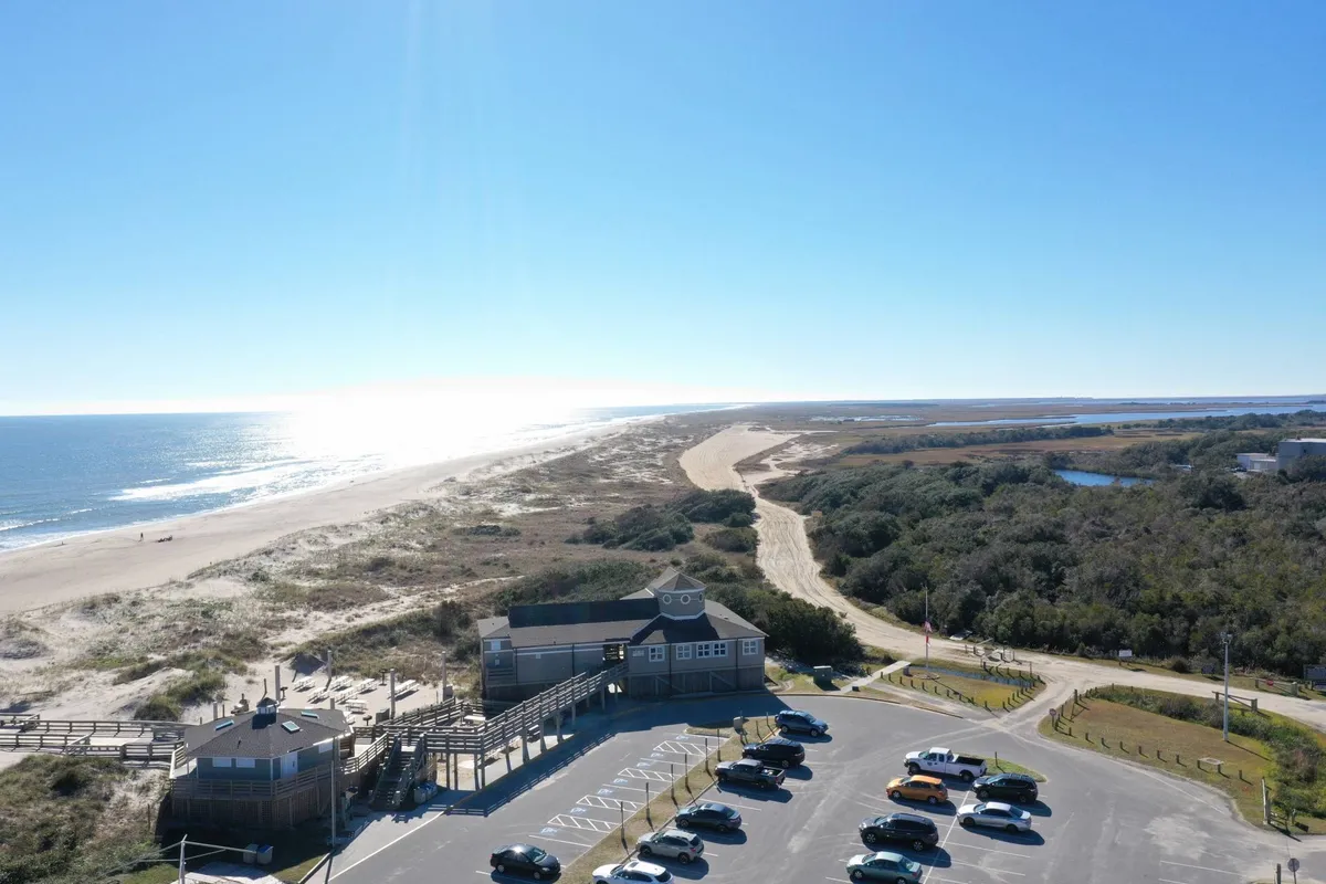 A wide exterior view featuring the sandy beach, gentle waves, nearby parking lot, and a glimpse of the Boardwalk. Lush coastal vegetation adds a natural touch, while the open space invites activities like swimming, surfing, and sunbathing.