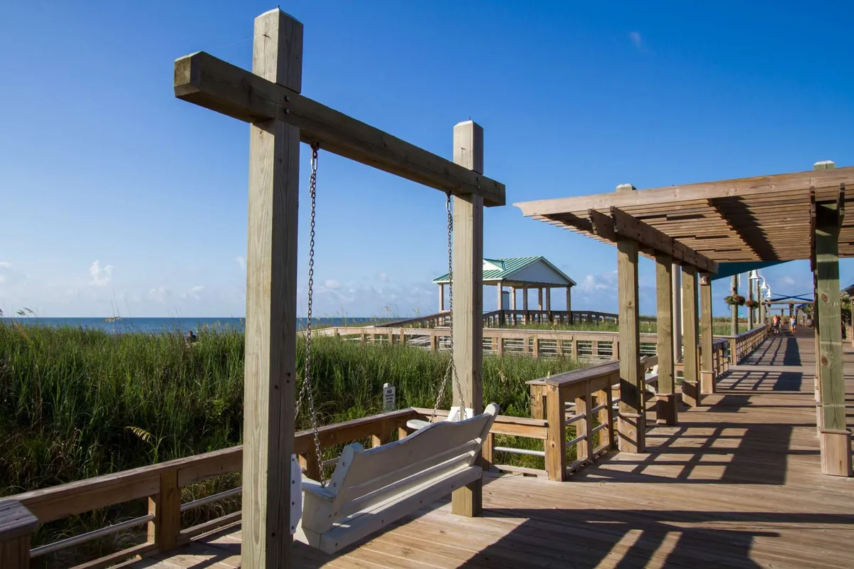 A tranquil scene featuring a swing on a boardwalk surrounded by green grass and pavilions, looking out toward the horizon. The atmosphere is serene, ideal for contemplative moments, sunset watching, or sharing time together in nature.