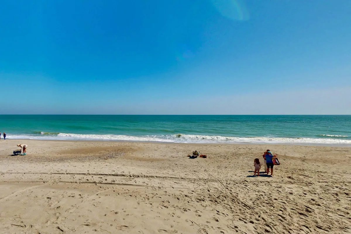 A cheerful beach scene showing people walking and playing on the sand, with the ocean waves in the background. The atmosphere is lively and family-oriented, perfect for beach games, sandcastles, and shell collecting.