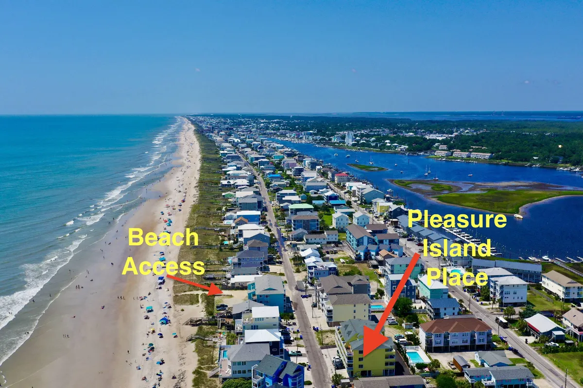 The hero image showcases Pleasure Island Place’s expansive balcony overlooking the sparkling ocean, easy beach access just steps away, tranquil residential homes, and the unique meeting point of ocean and inland waterways. The inviting, sun-drenched scene highlights leisurely vacation activities like morning sunrises, strolls along sandy shores, swimming, and exploring the vibrant coastal community.