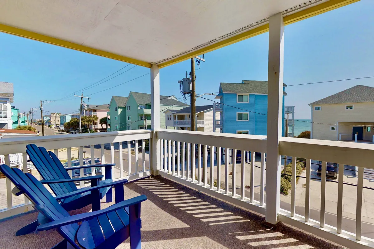 A pair of blue Adirondack chairs set on a porch or balcony overlook neighboring houses with the ocean visible in the distance. The scene evokes relaxation and quiet moments, perfect for enjoying morning coffee or watching the sunset together.