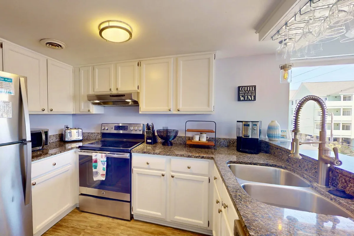 The kitchen is the focal point of this image, showcasing sleek stainless steel appliances, a granite countertop, a kitchen island with a sink, and accents like a 'coffee and wine' sign and hanging glasses. The space is functional and welcoming, with ample prep area and elements that evoke gatherings and culinary rituals.