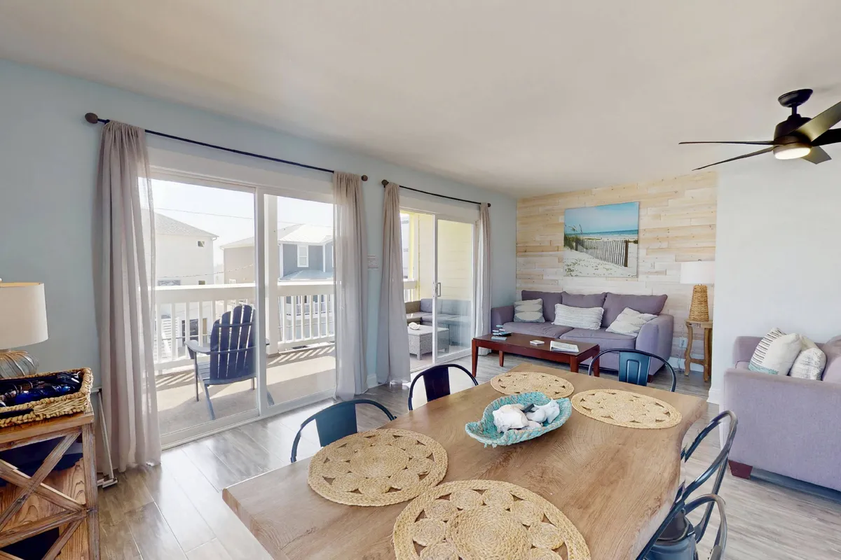 A welcoming, sunlit interior showing a dining table near sliding glass doors, a comfortable sofa with cushions, and tasteful wall art. The area exudes warmth and is designed for both casual relaxation and shared meals, with ample natural light enhancing the homely feel.