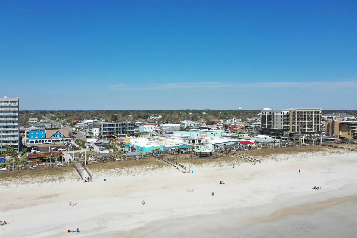 A lively stretch of Carolina Beach with colorful buildings lining the shore, piers jutting into the water, and people relaxing and socializing on the sandy beach. The scene captures local shops, restaurants, and entertainment spots, all within view of the ocean.