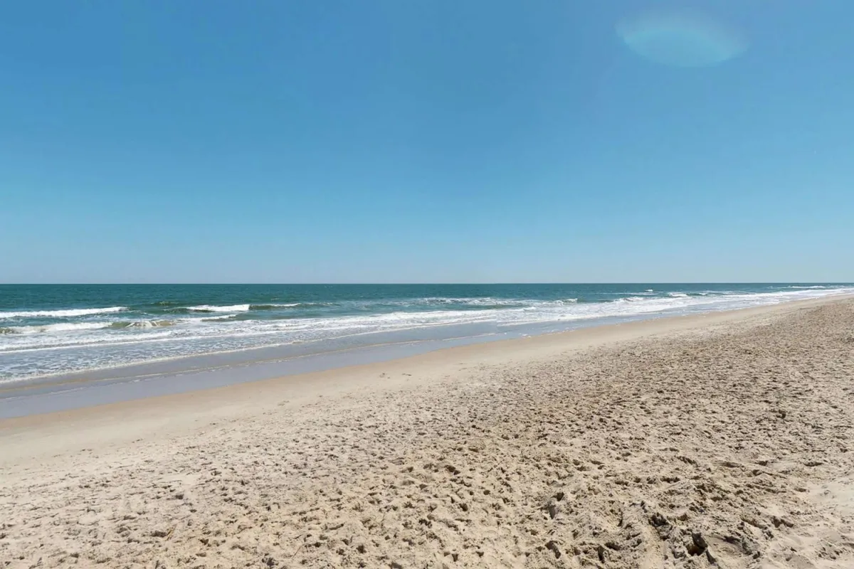A serene stretch of sandy beach with gentle ocean waves under a clear blue sky. The inviting shoreline is perfect for sunbathing, morning walks, or collecting seashells.