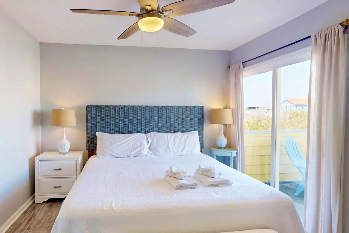 A tranquil bedroom scene featuring a bed with crisp white linens, a stylish blue headboard, and nightstands with lamps on either side. A sliding glass door opens to a balcony, inviting in natural light and offering a peaceful view.