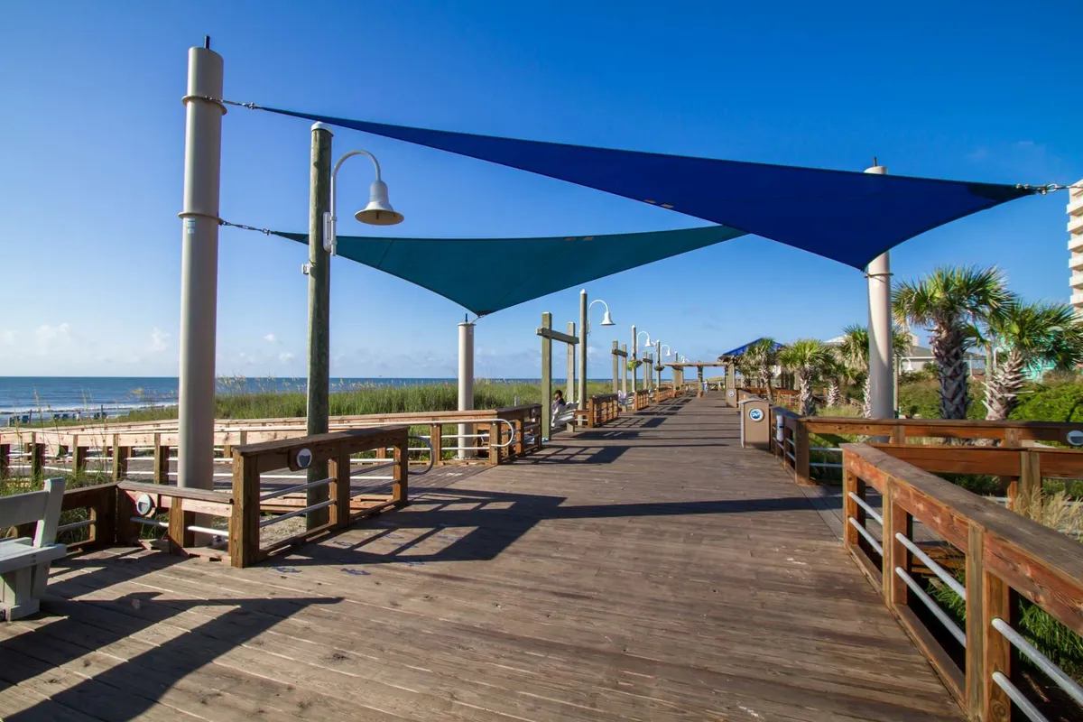 A scenic exterior shot of a wooden boardwalk stretching toward the ocean, accented with blue shade sails, palm trees, benches, and a sweeping ocean view. The setting invites visitors to stroll, relax, or enjoy the coastal breeze.